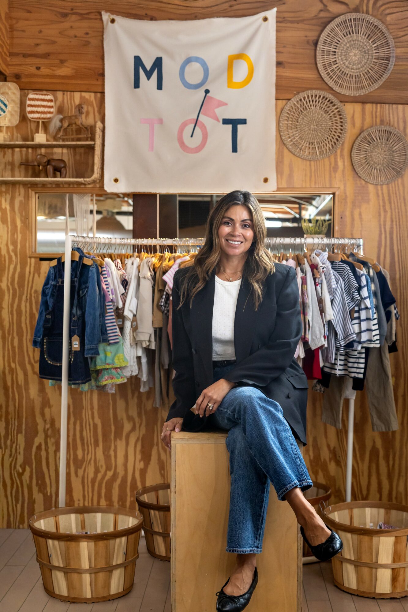 Woman sitting on a wooden block in a clothing store, smiling, with racks of clothes behind her and decorative baskets on the wall.