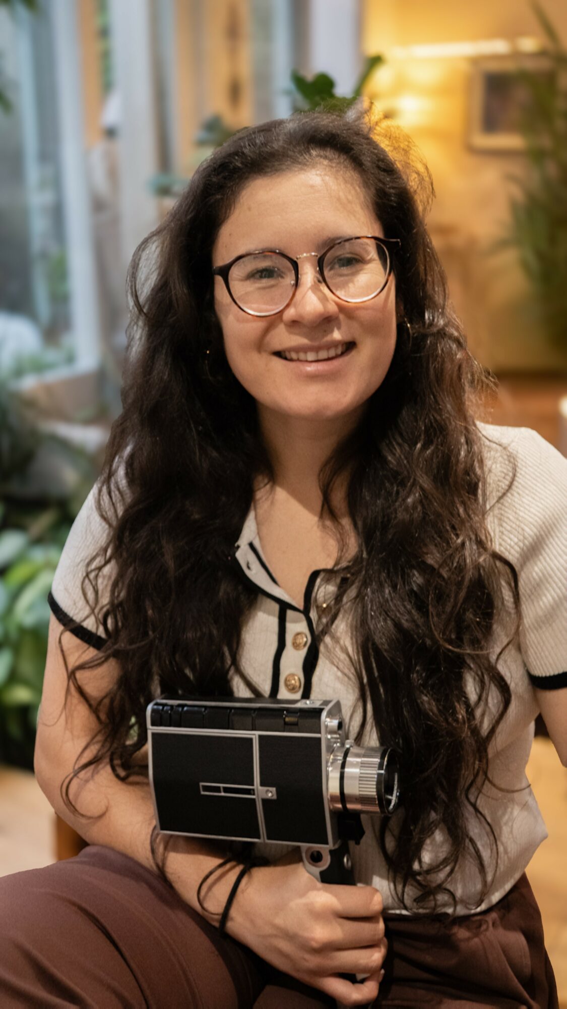 Woman with long dark hair and glasses smiling indoors, holding a camera or device, in a cozy room with plants and warm lighting.