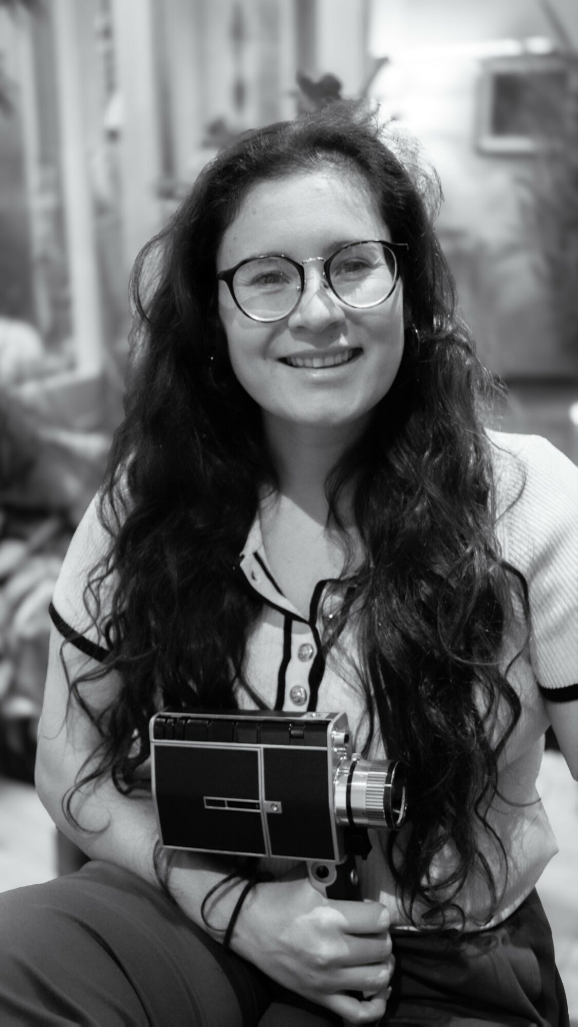 Young woman with glasses and long curly hair smiling, holding a smartphone, in an indoor setting.