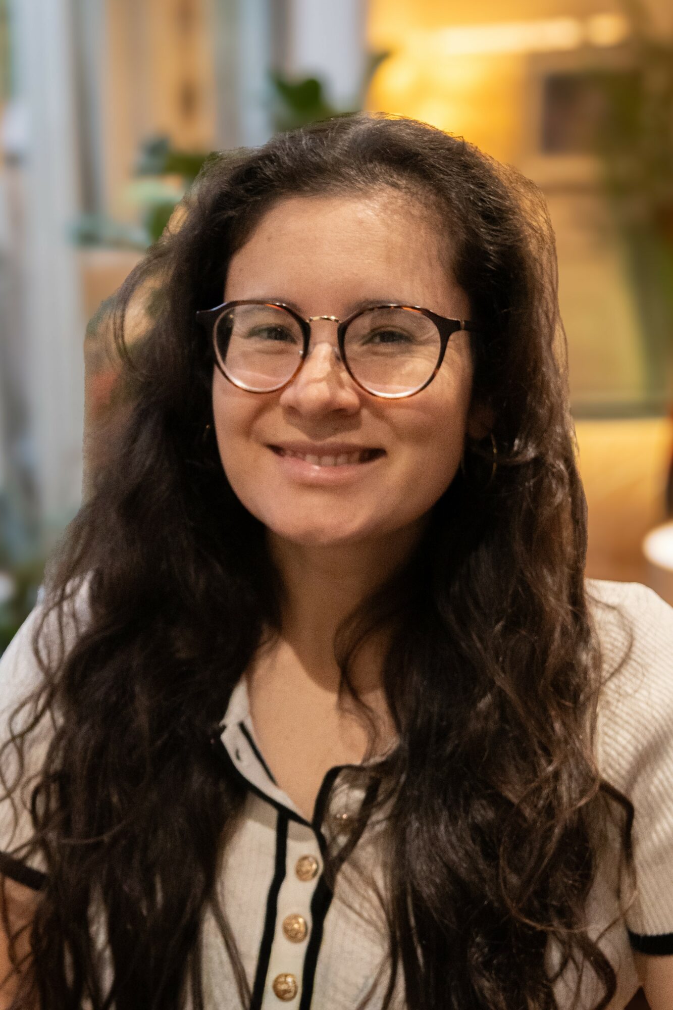 Young woman with long dark hair, glasses, smiling, indoors with warm lighting and plants in background.