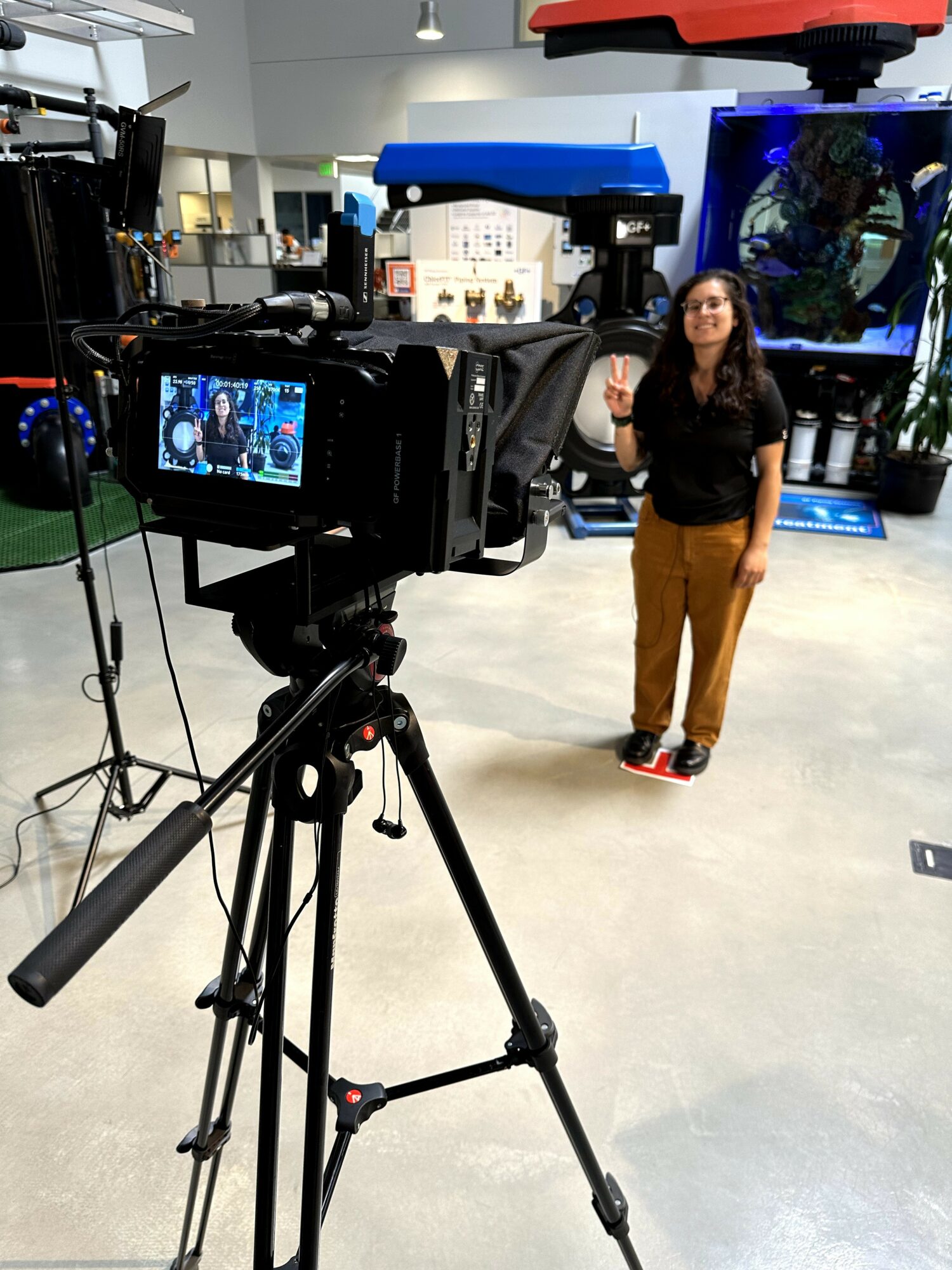 Woman standing in front of a large screen, being filmed in a studio setting, with camera equipment visible.