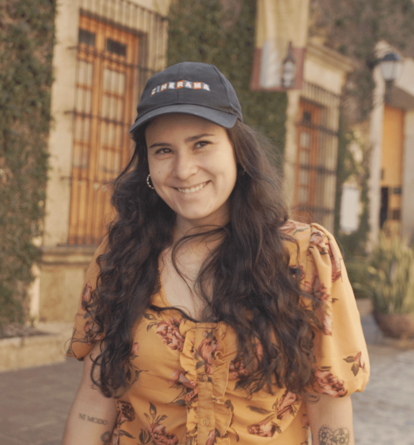 Young woman with long dark hair wearing a black cap and floral yellow top, smiling outdoors.