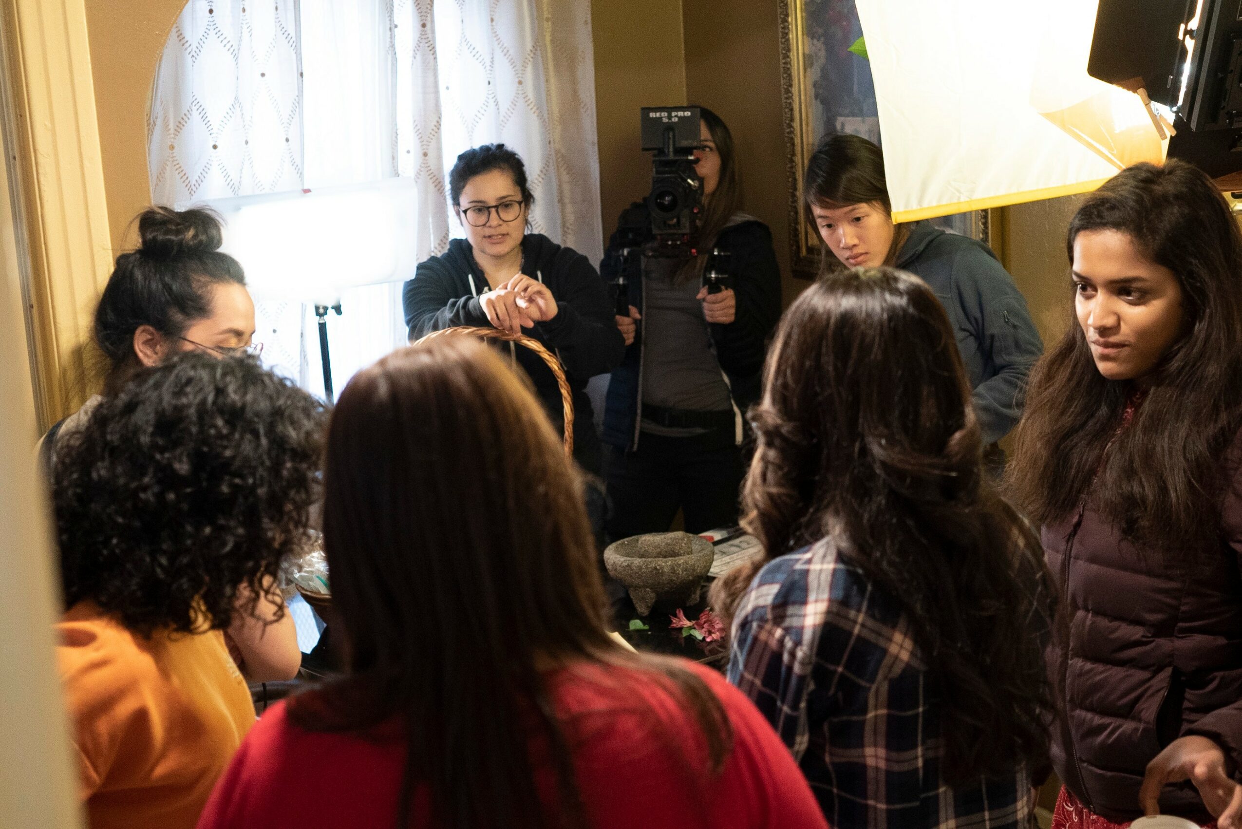 Group of people gathered indoors, some speaking and others listening, with a camera and lighting equipment present.