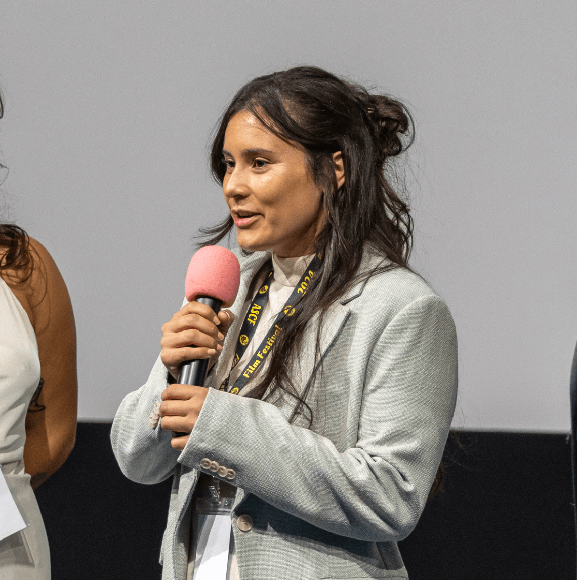 Woman with long dark hair speaking into a pink microphone, wearing a light gray blazer, in front of a plain background.