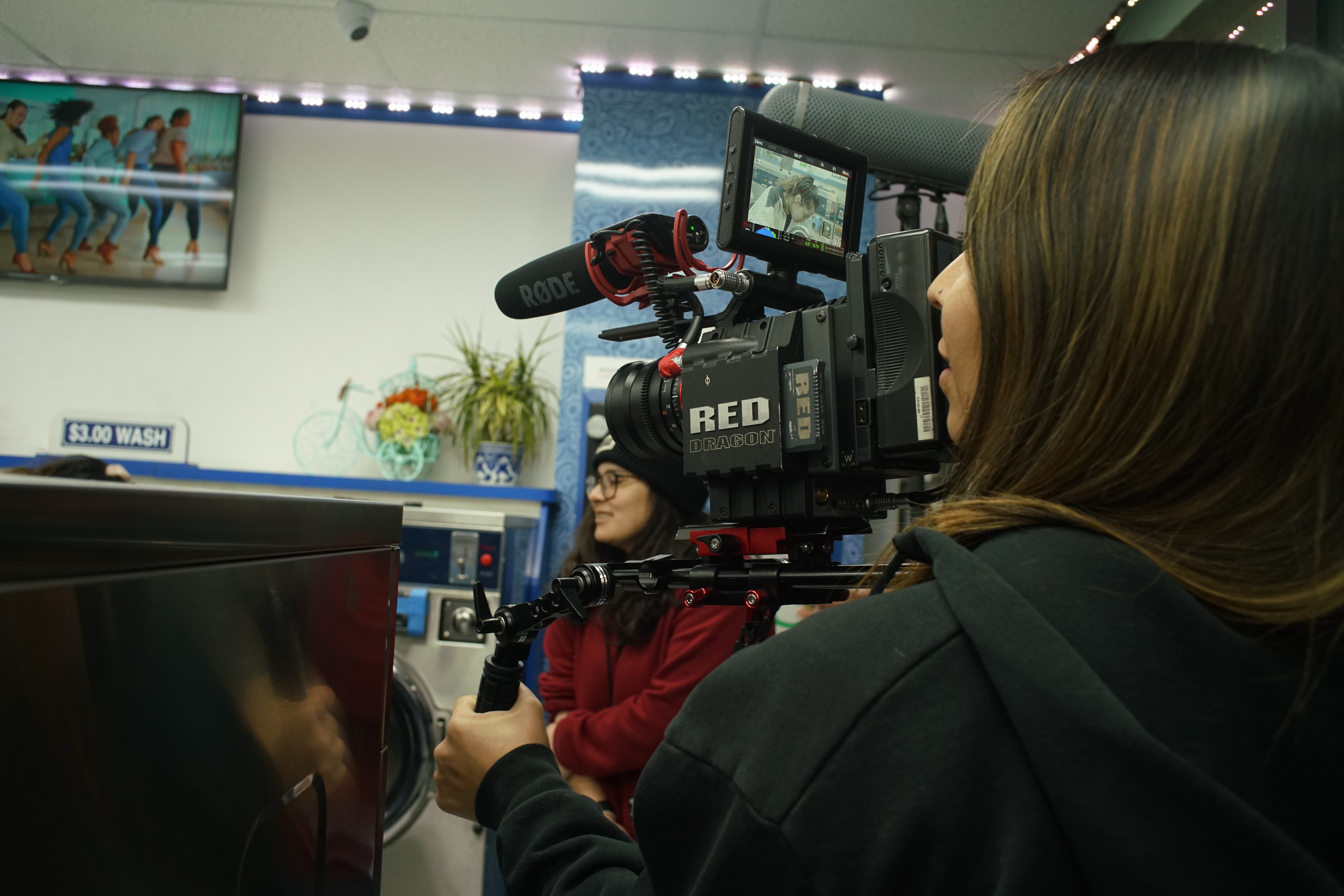 Person operating a professional video camera indoors, with another person in the background, and a television screen on the wall.