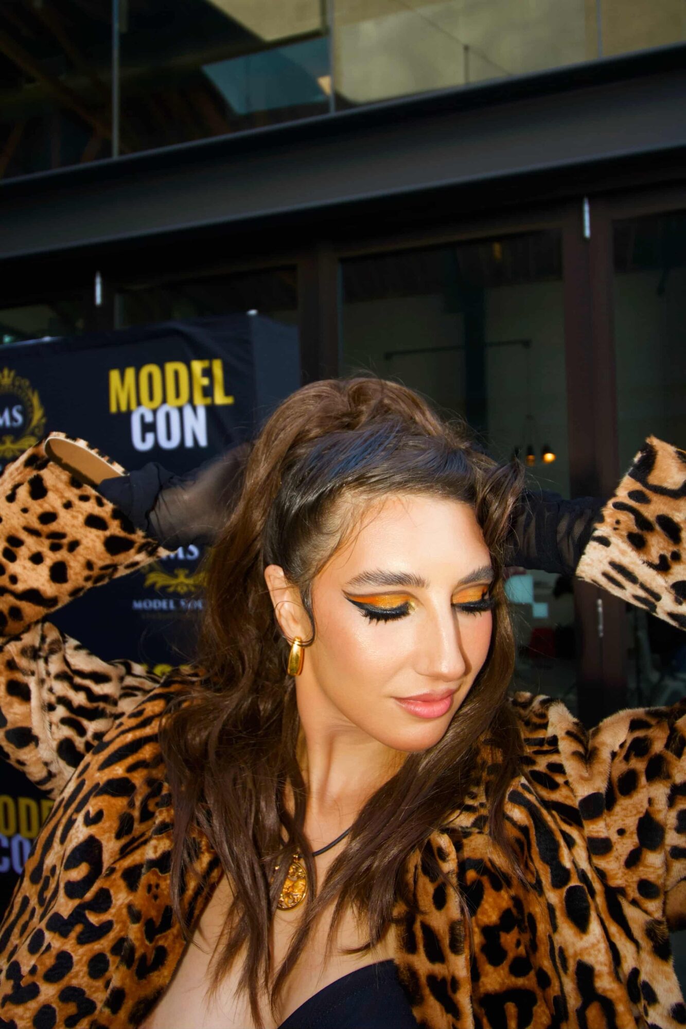 Woman with long brown hair wearing leopard print top, eyes closed, adjusting her hair with both hands, in front of a glass building.