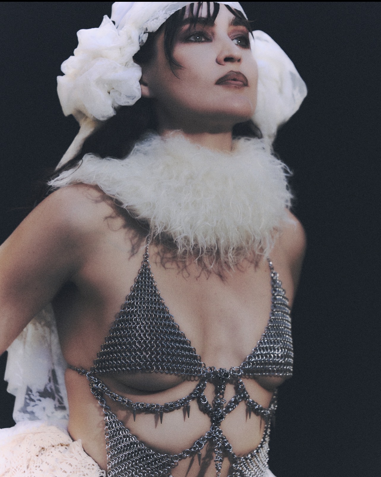 Woman wearing a headpiece and a beaded top with decorative chains, posing against a dark background.