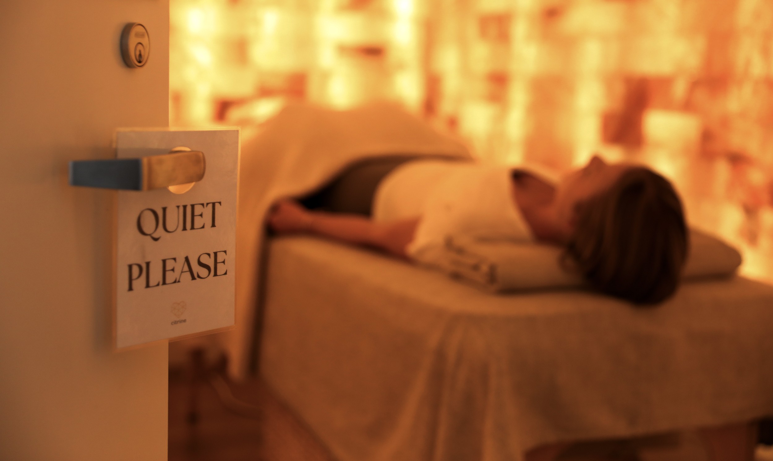 Person lying on bed with head turned away, in a warmly lit room, door with a sign reading 'Quiet Please'.