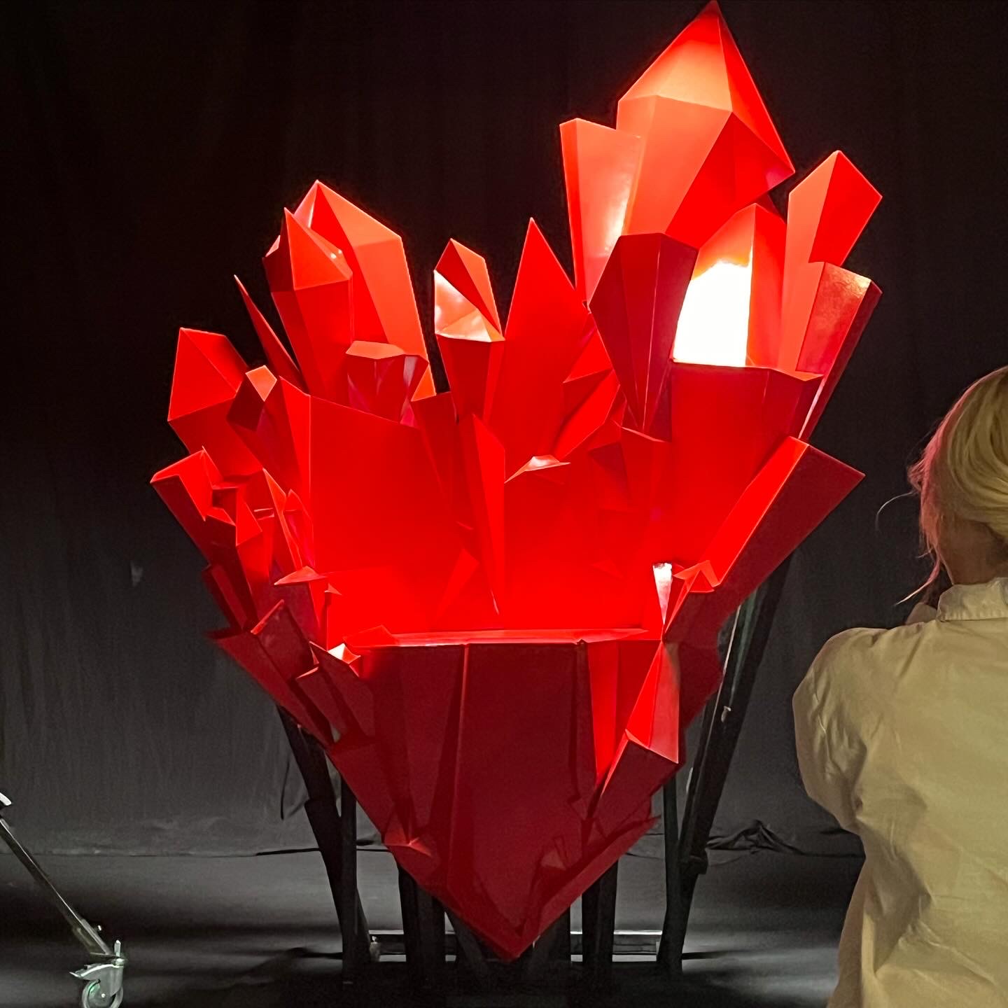 Large red geometric sculpture resembling a heart, displayed indoors with a person in a yellow jacket nearby.