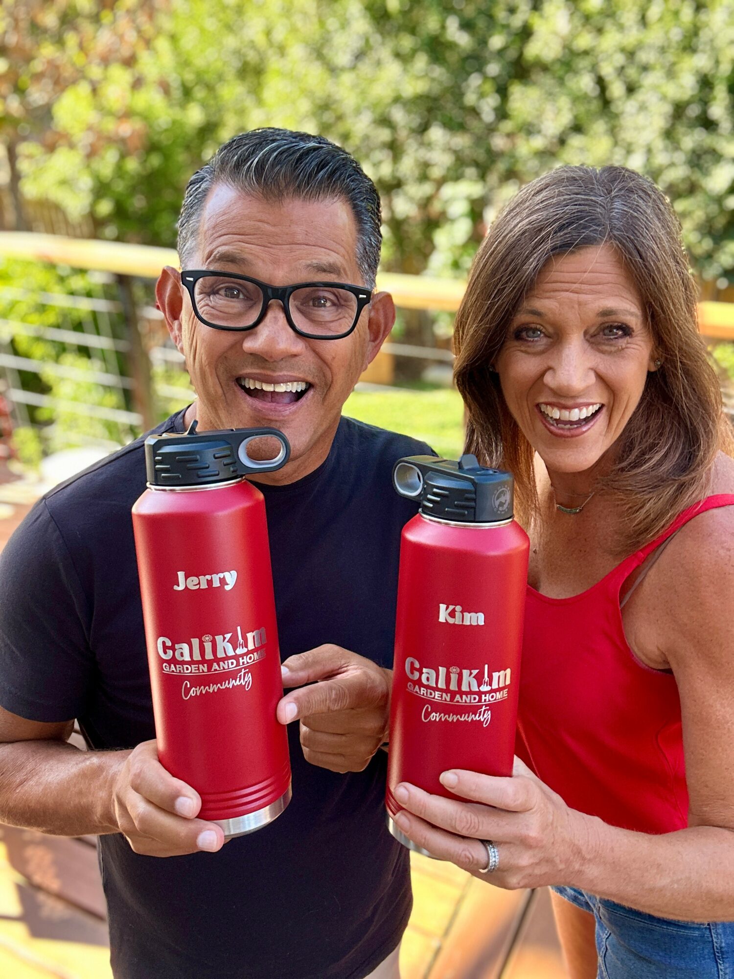 Two smiling people holding red water bottles outdoors with trees in the background.
