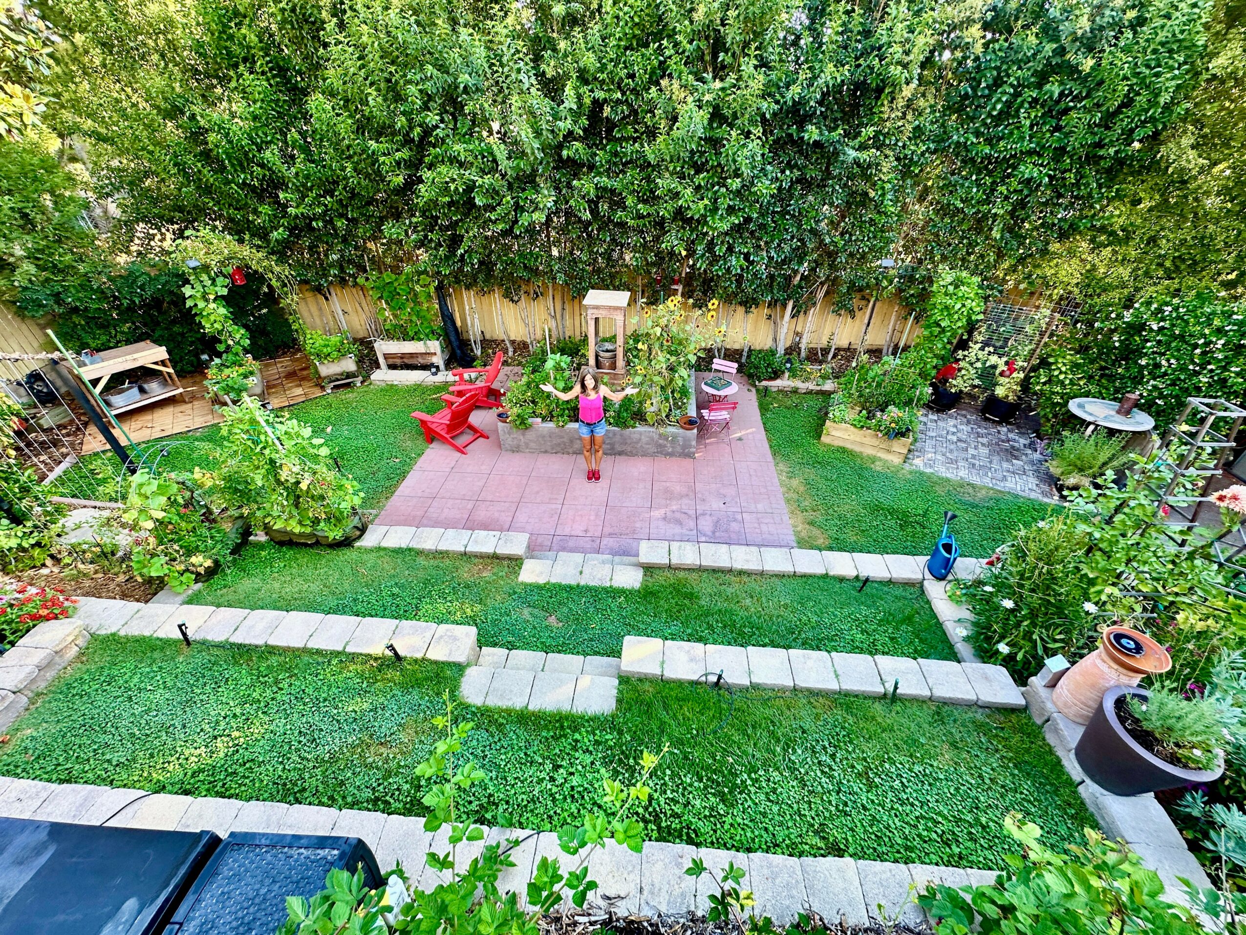 Backyard garden with patio, red chairs, green grass, trees, and potted plants, viewed from above.