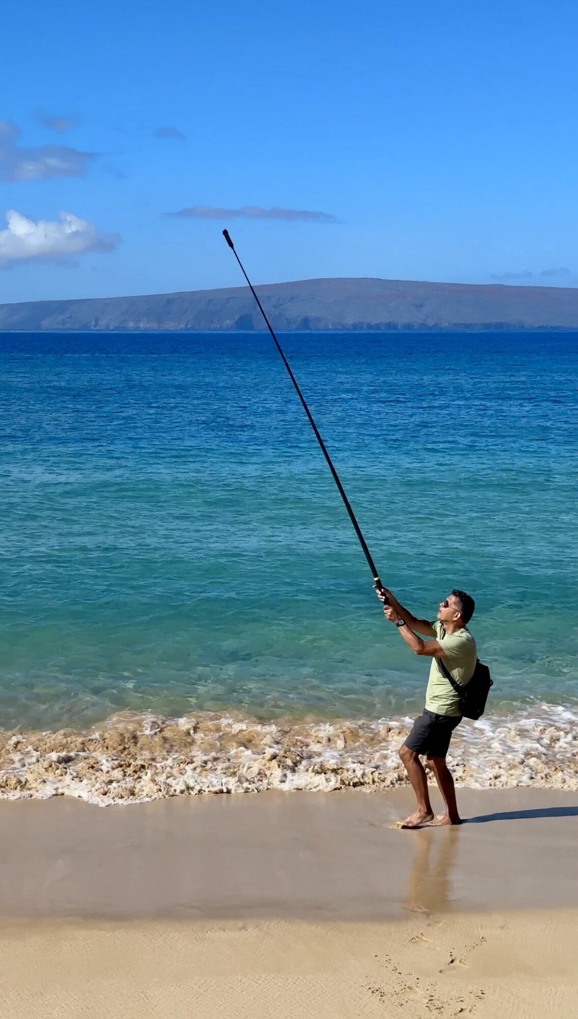 Child standing on beach holding a long fishing pole over water, with ocean and sky in background.