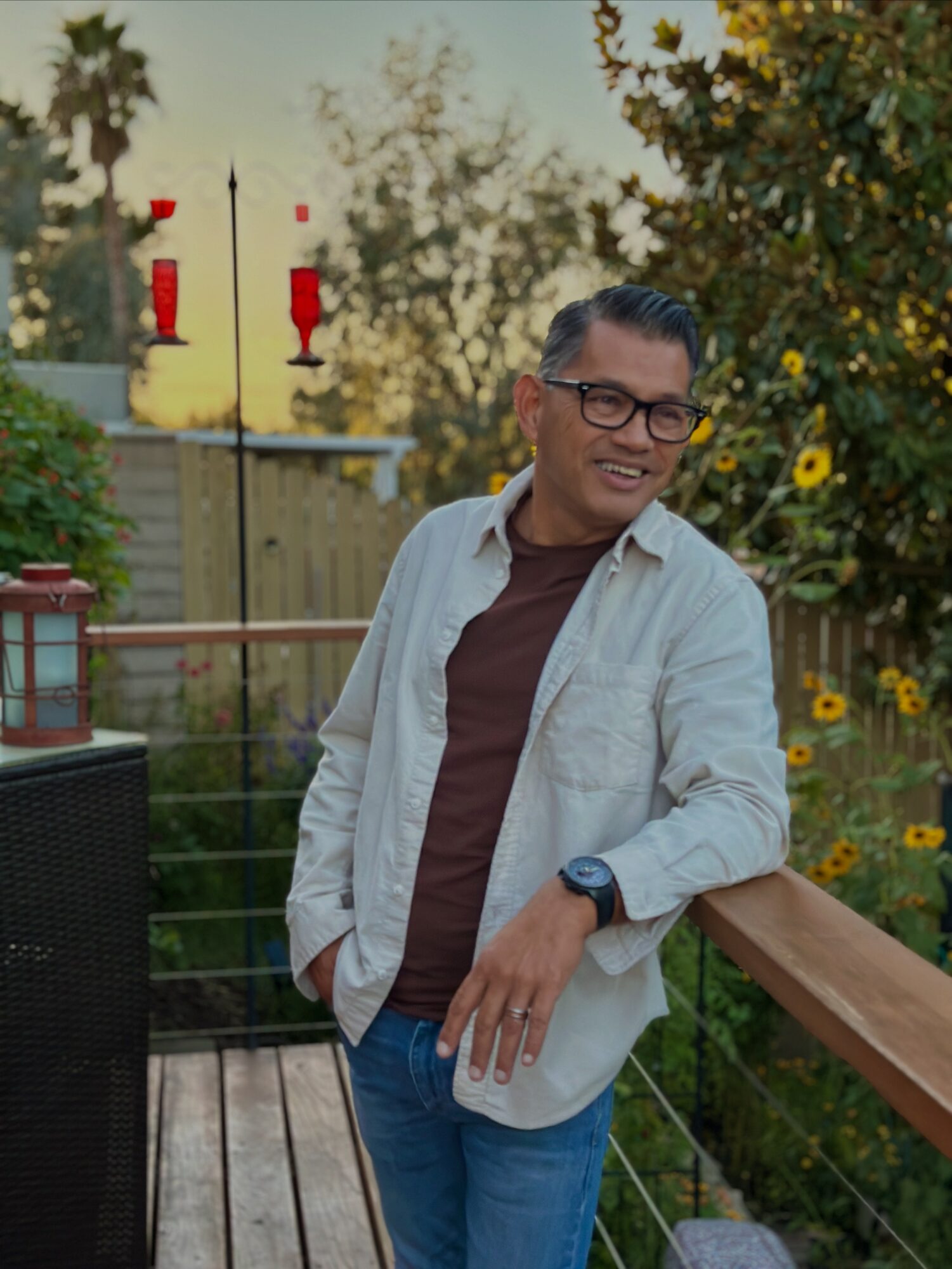 Man with glasses and a watch leaning on a wooden railing outdoors, surrounded by trees and flowers, during daytime.