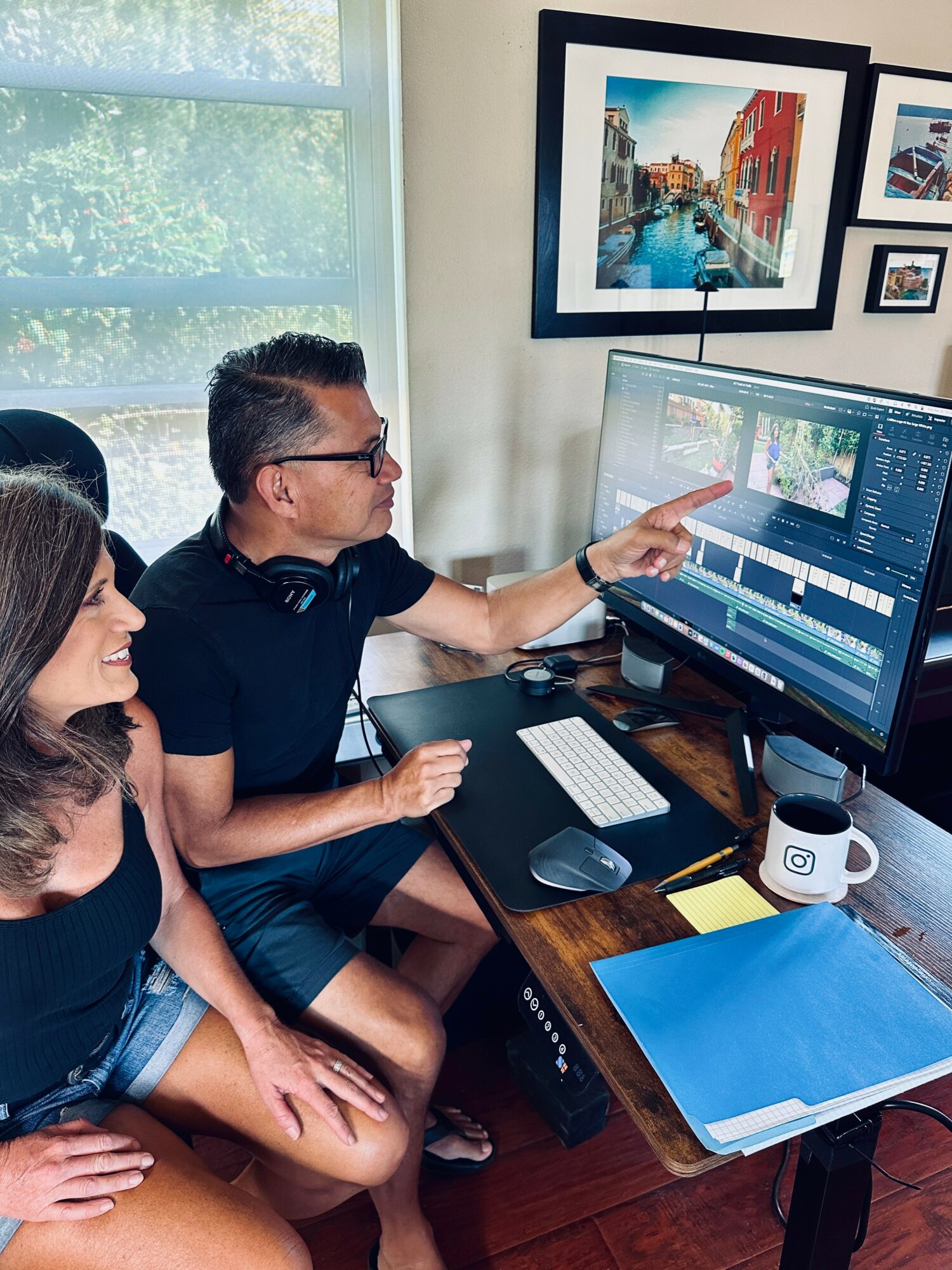 Two people working on video editing at a desk with a computer, framed pictures on the wall, and a window in the background.
