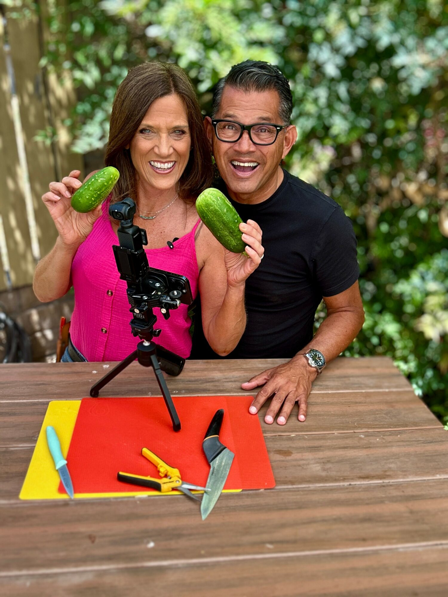 Two people smiling and holding cucumbers outdoors, with gardening tools and a camera on a table.