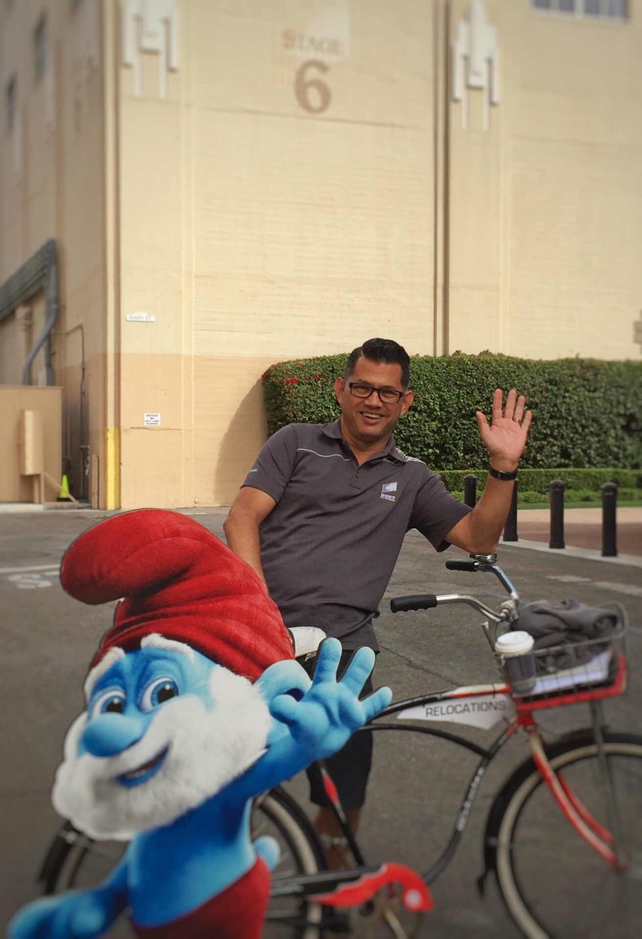 Man waving while standing next to a bicycle and a Smurf mascot in front of a building with a hedge.