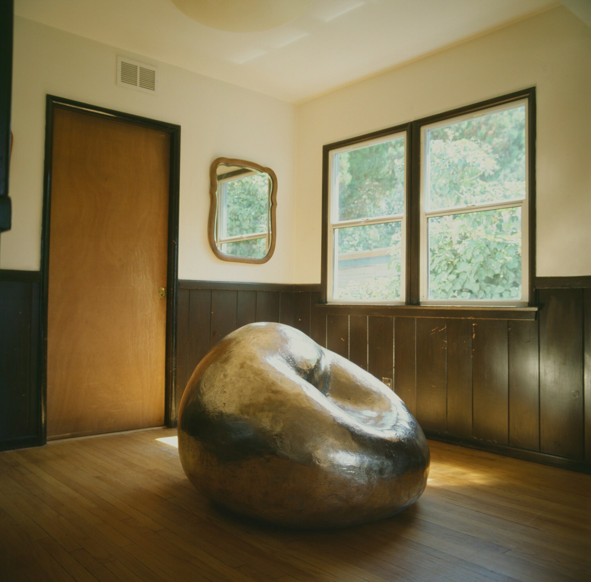 Room with wooden floor, two windows, a door, a mirror, and a large metallic sculpture resembling a rounded stone.