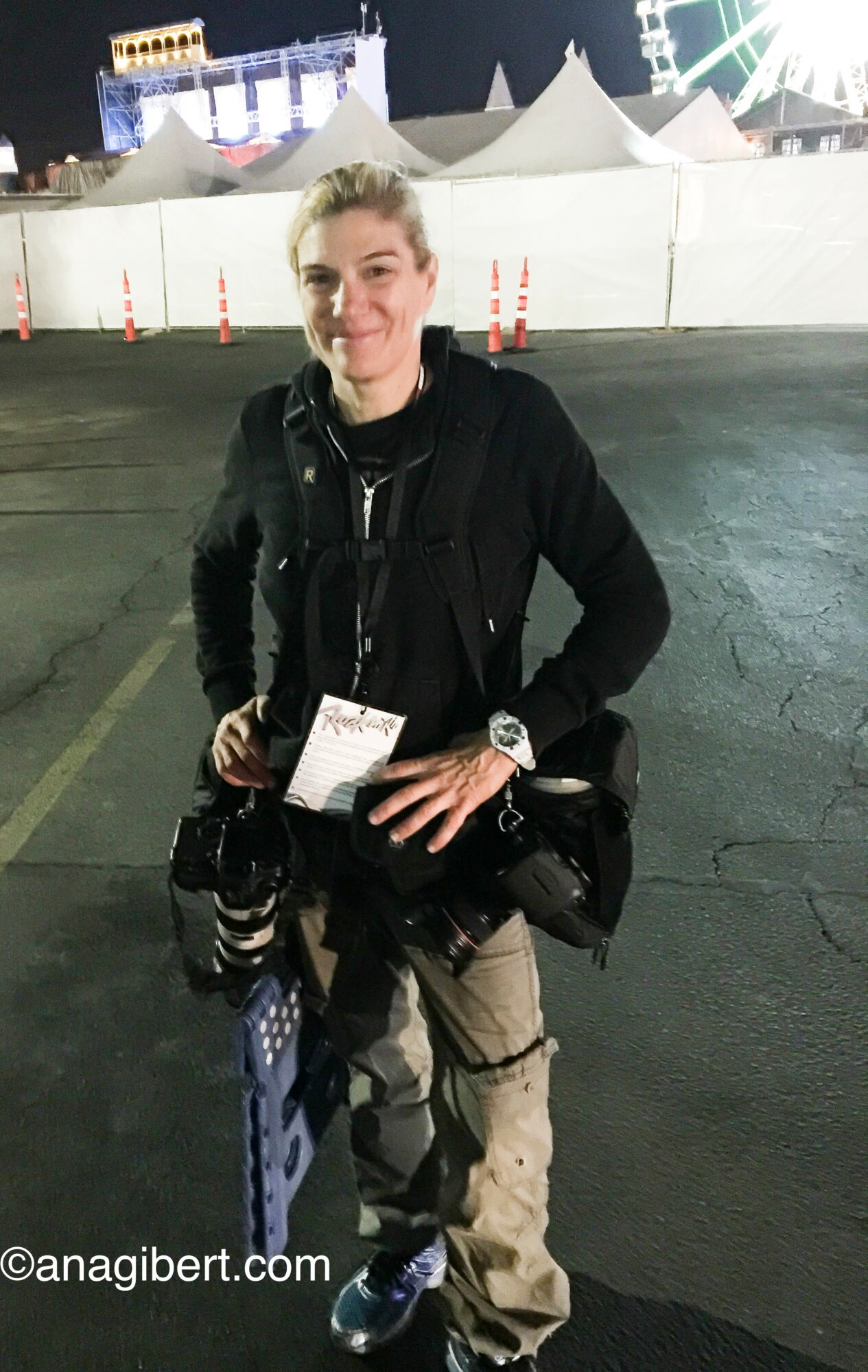 Young person with short hair smiling, standing outdoors at night with tents and lights in background, holding a ticket and camera gear.