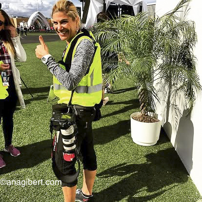 Woman in a yellow safety vest smiling and giving a thumbs-up outdoors with a potted plant nearby.