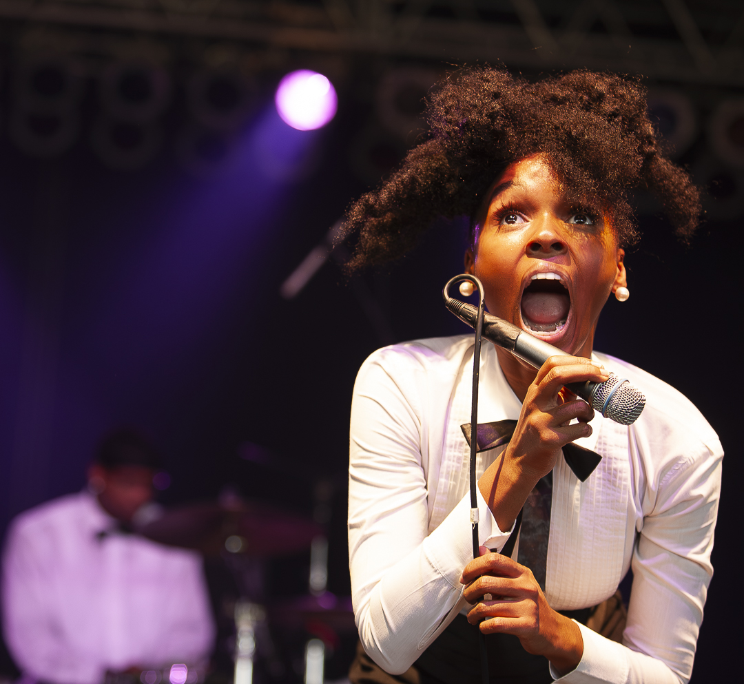 A woman singing passionately into a microphone on stage with a band in the background, stage lights shining down.