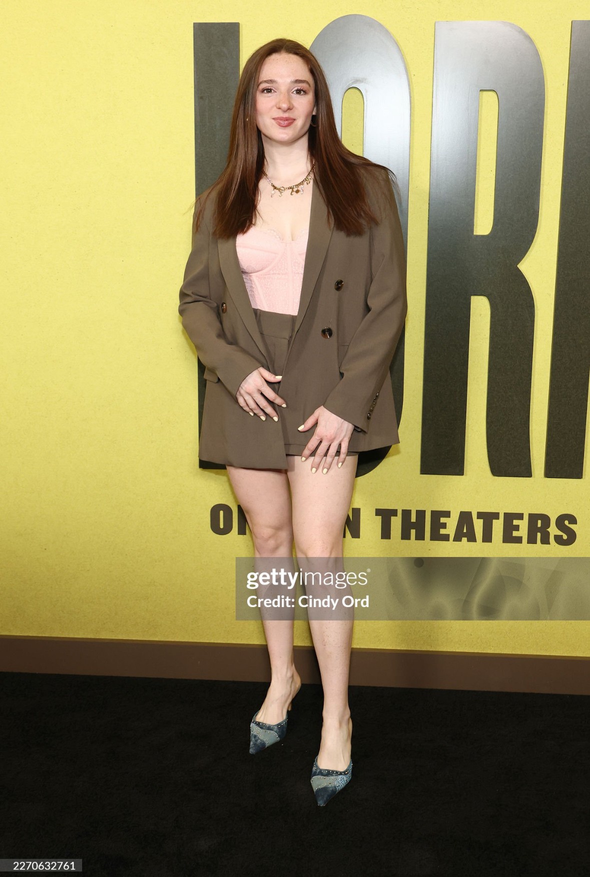 Young woman with long brown hair in a blazer and dress, standing in front of a yellow background with text.