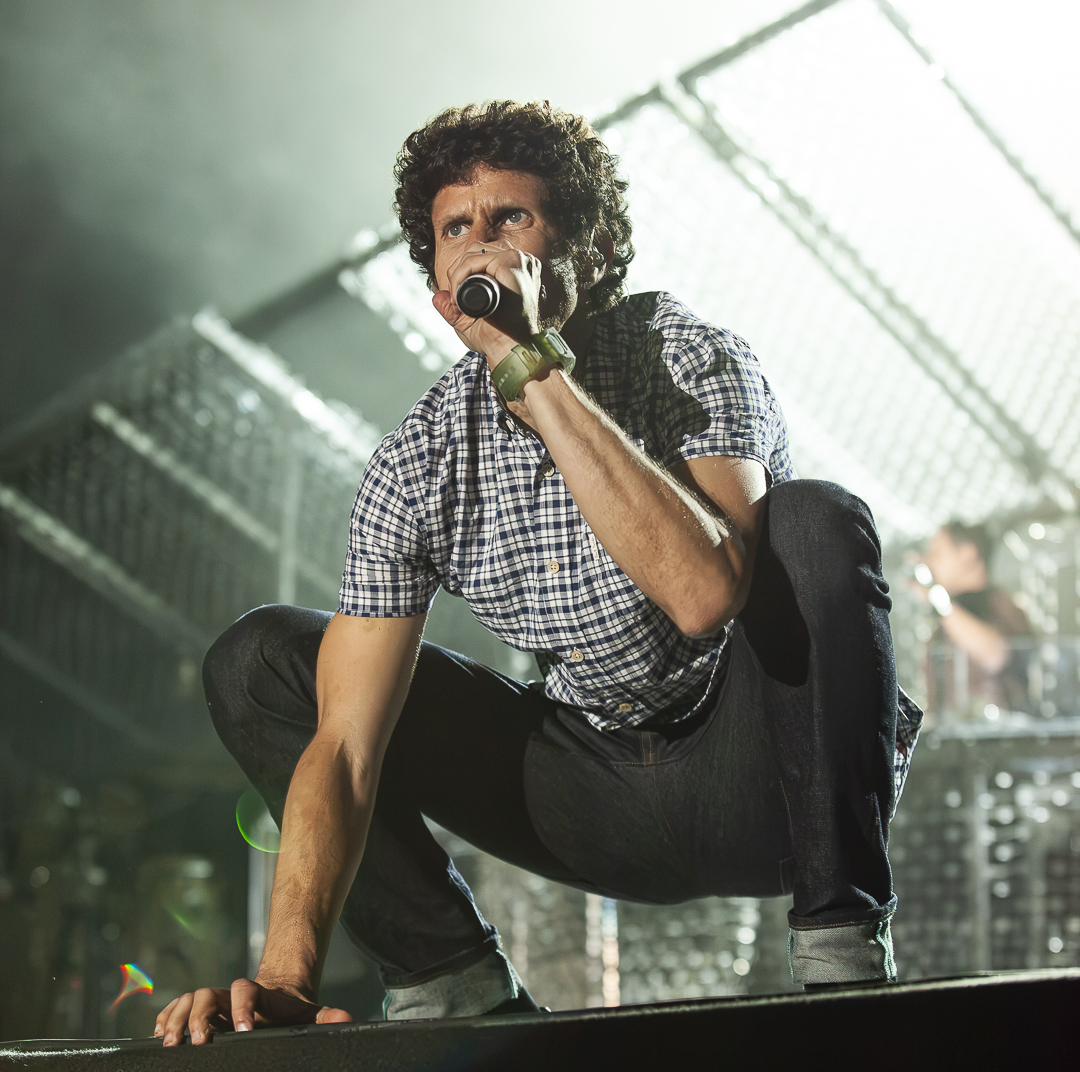 Performer crouching on stage holding microphone, wearing checkered shirt, with stage lights and equipment in background.