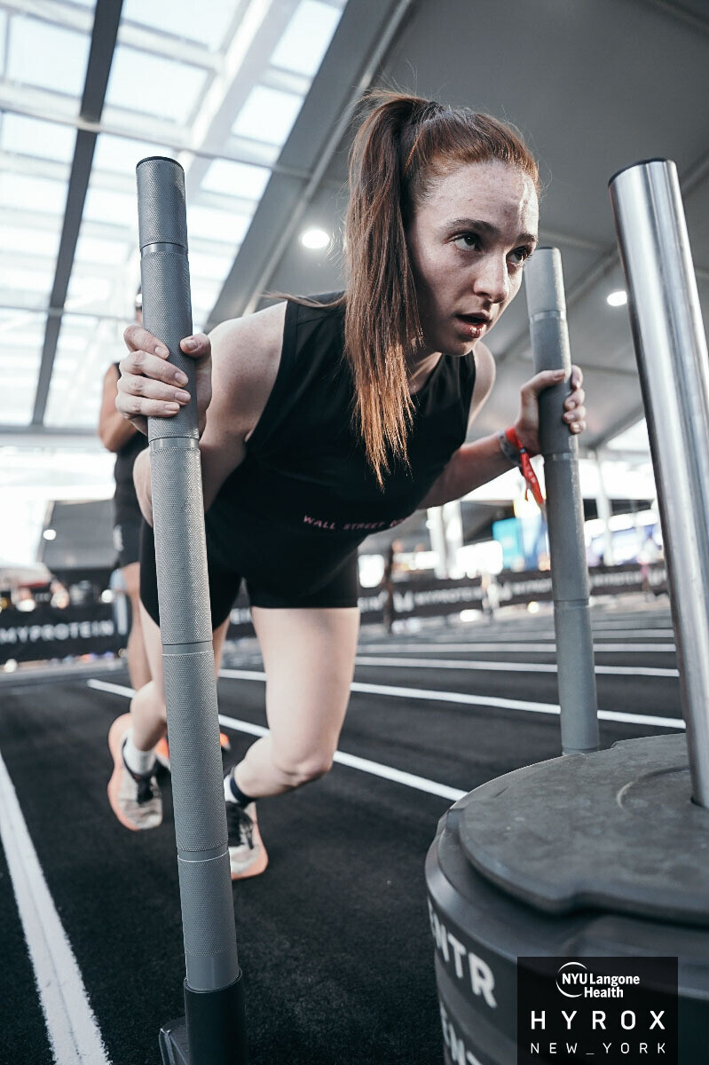 Young woman in workout clothes pushes a sled at gym, focused on exercise, with large windows in background.