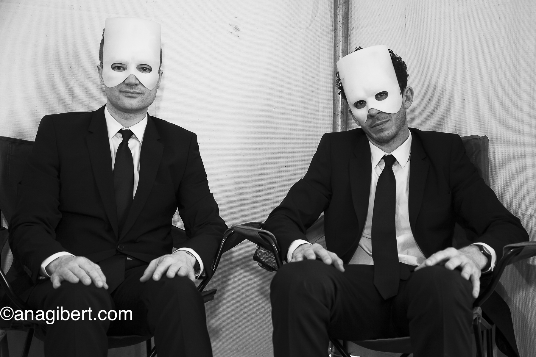Two people in suits wearing white masks sit in chairs against a plain background.