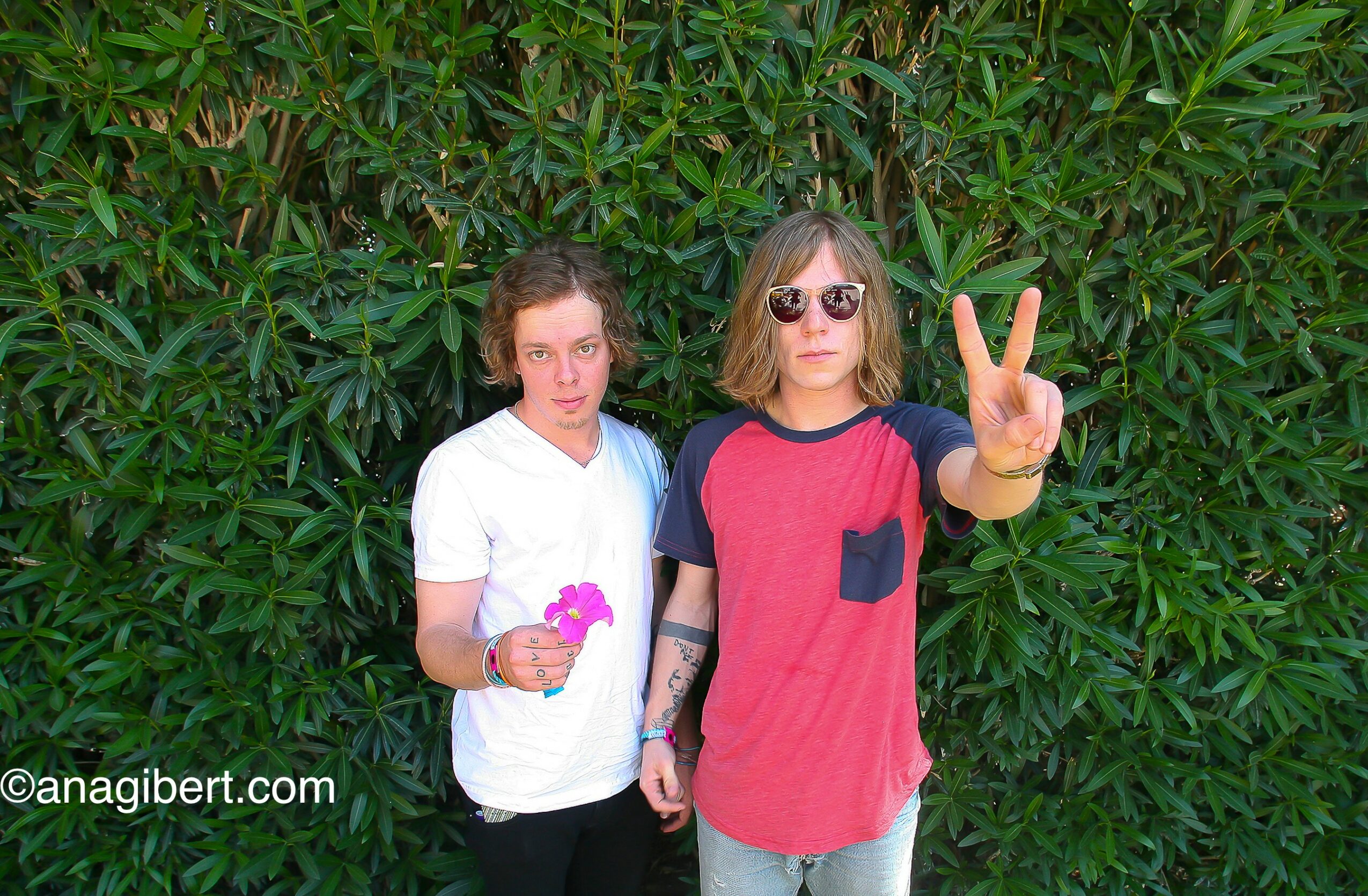 Two women standing in front of green foliage, one making a peace sign with her hand, both wearing casual clothing and sunglasses.
