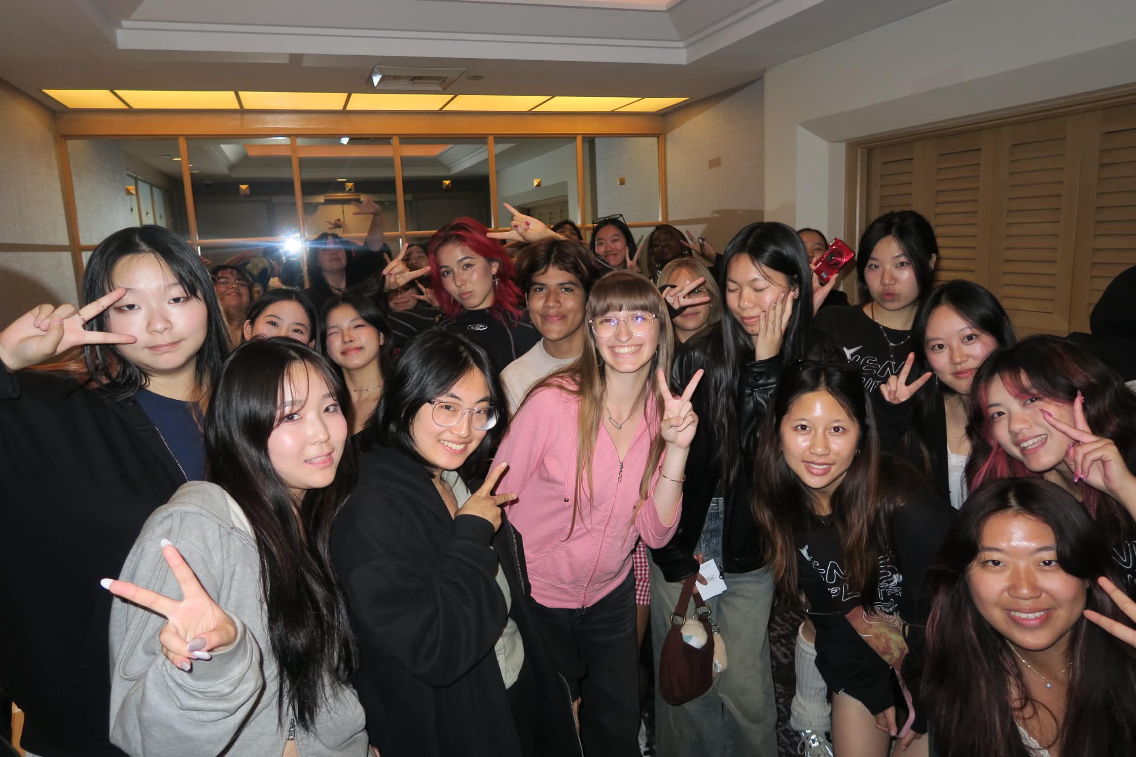 Group of young women and girls smiling and making peace signs indoors, some wearing glasses and casual clothing.