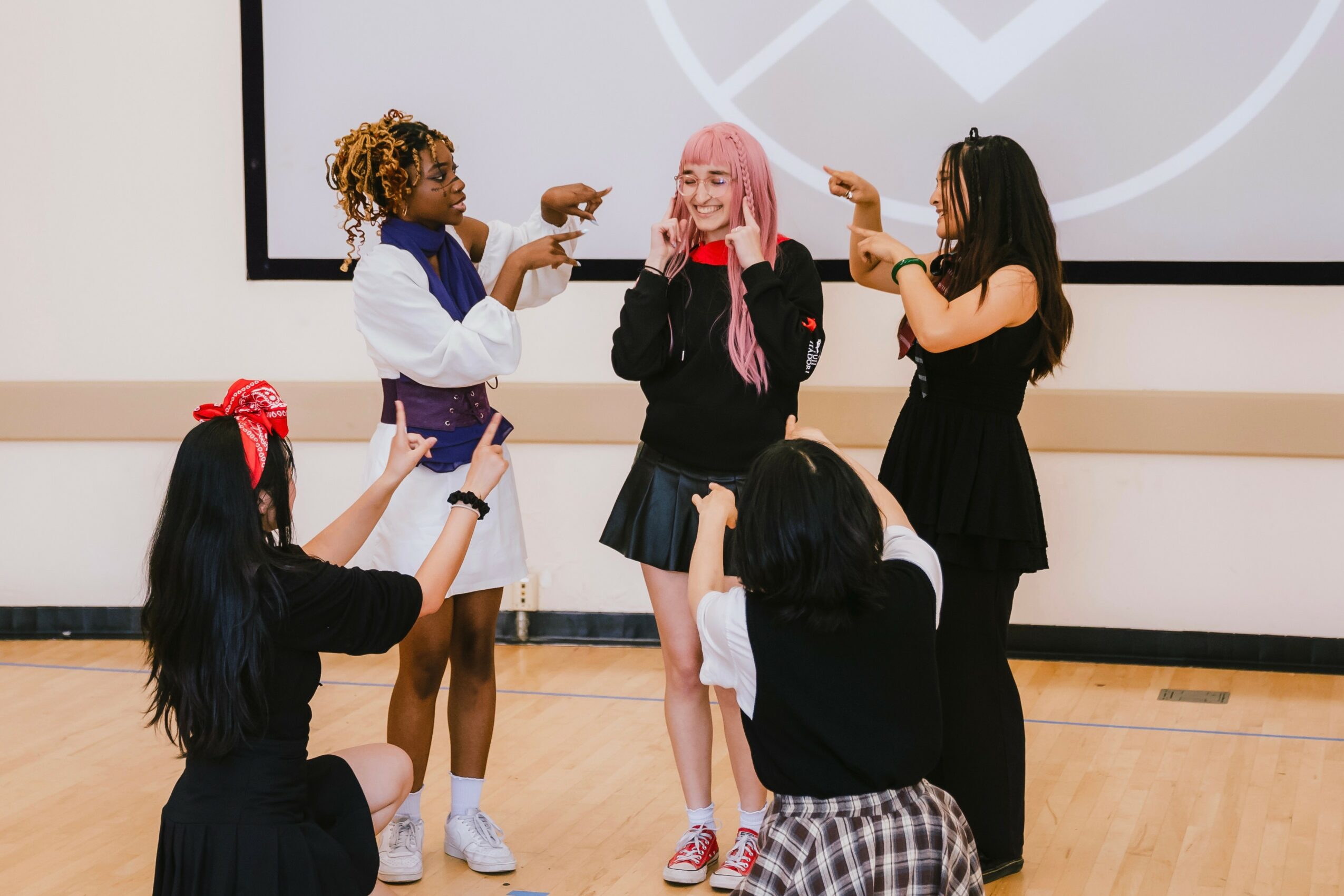 Five women, one with pink hair, are on stage, smiling and pointing at each other, with a large screen behind them.