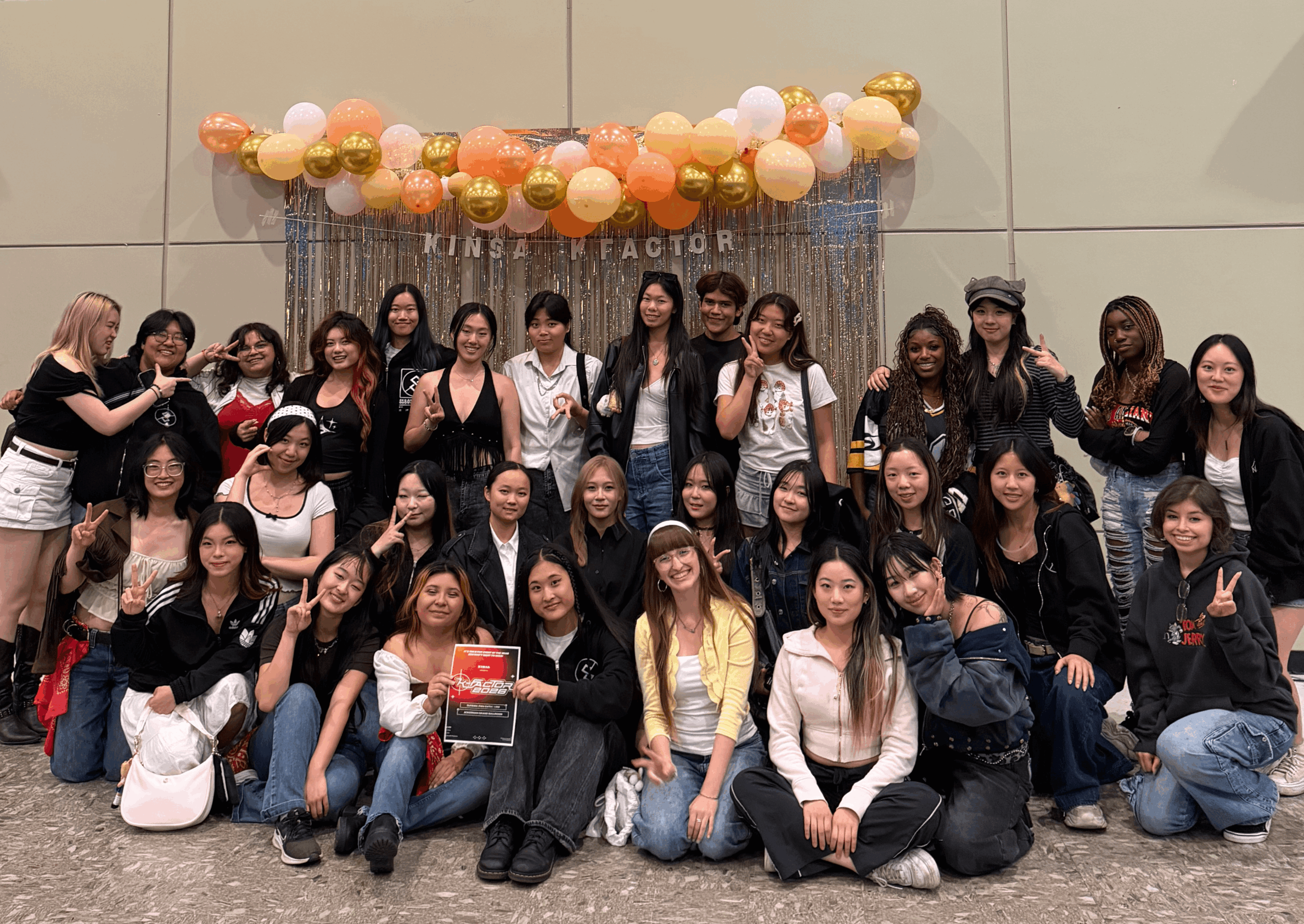 Group of women posing in front of a decorated backdrop with balloons, some making peace signs, smiling, and wearing casual clothing.