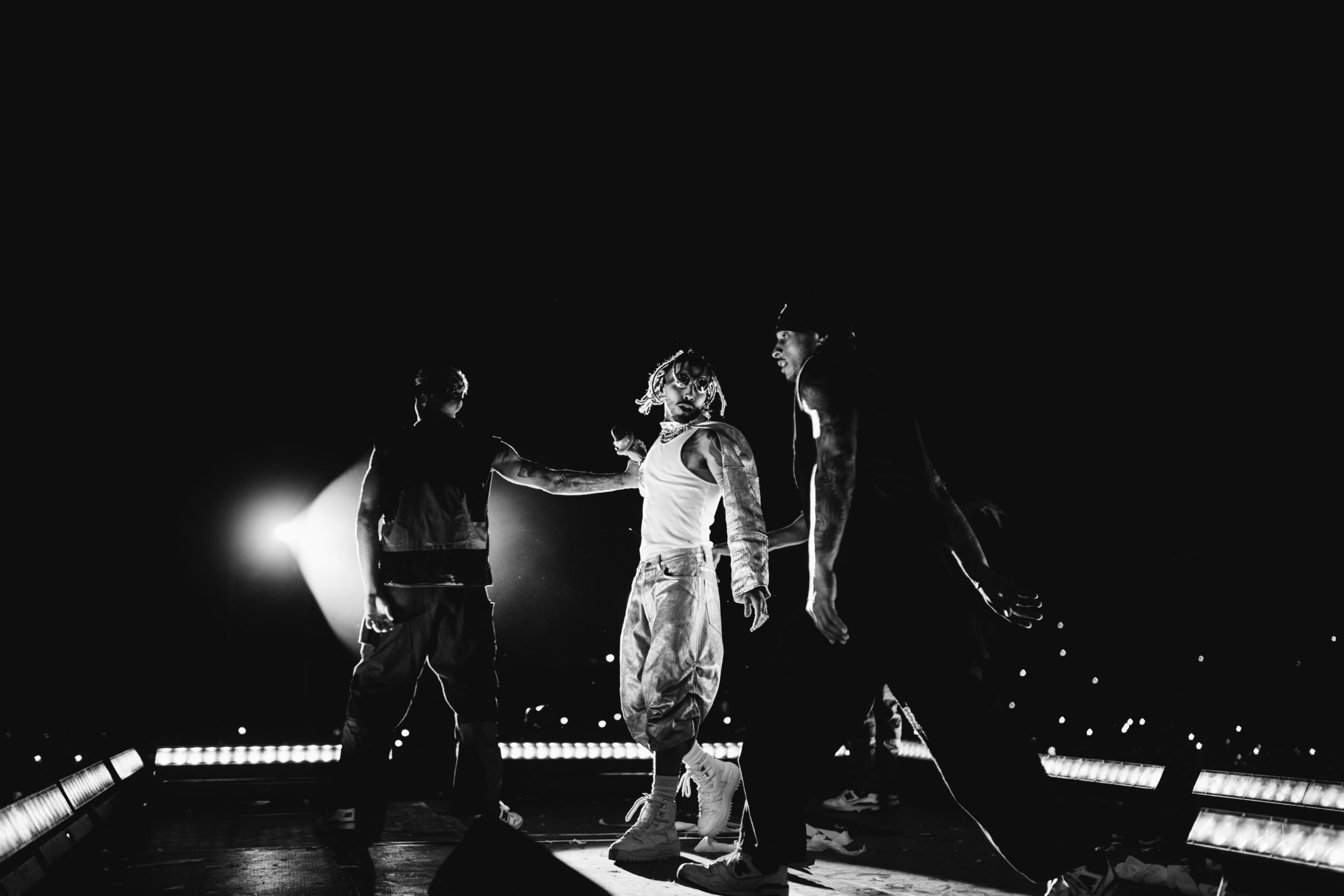 Three people standing on a stage with bright lights behind them, holding hands or connecting, in a dark setting.