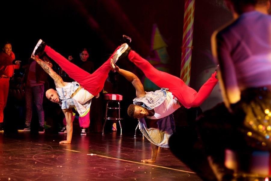 Two dancers perform handstands on stage with a group watching in the background, colorful lighting, and a festive atmosphere.