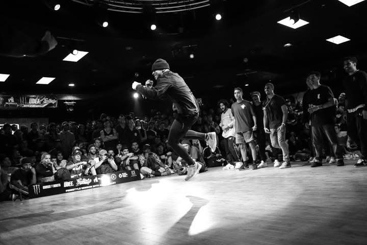 Person performing a dance move on stage with a crowd watching, in a dark indoor venue with overhead lights.