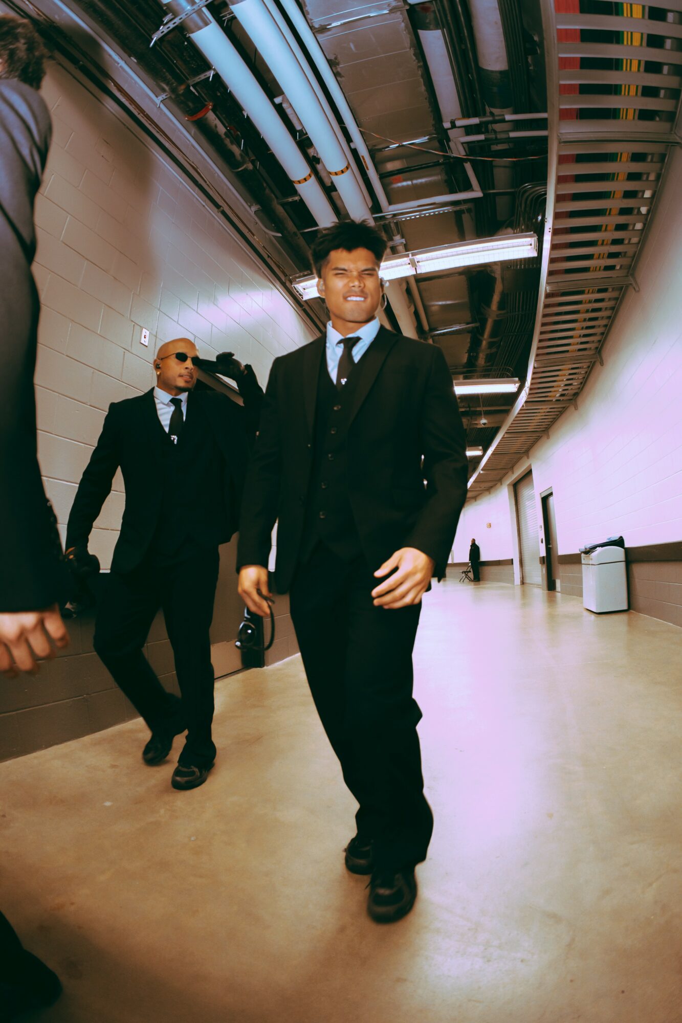 Two men in black suits and sunglasses walking in an indoor space with a curved ceiling and bright lighting.