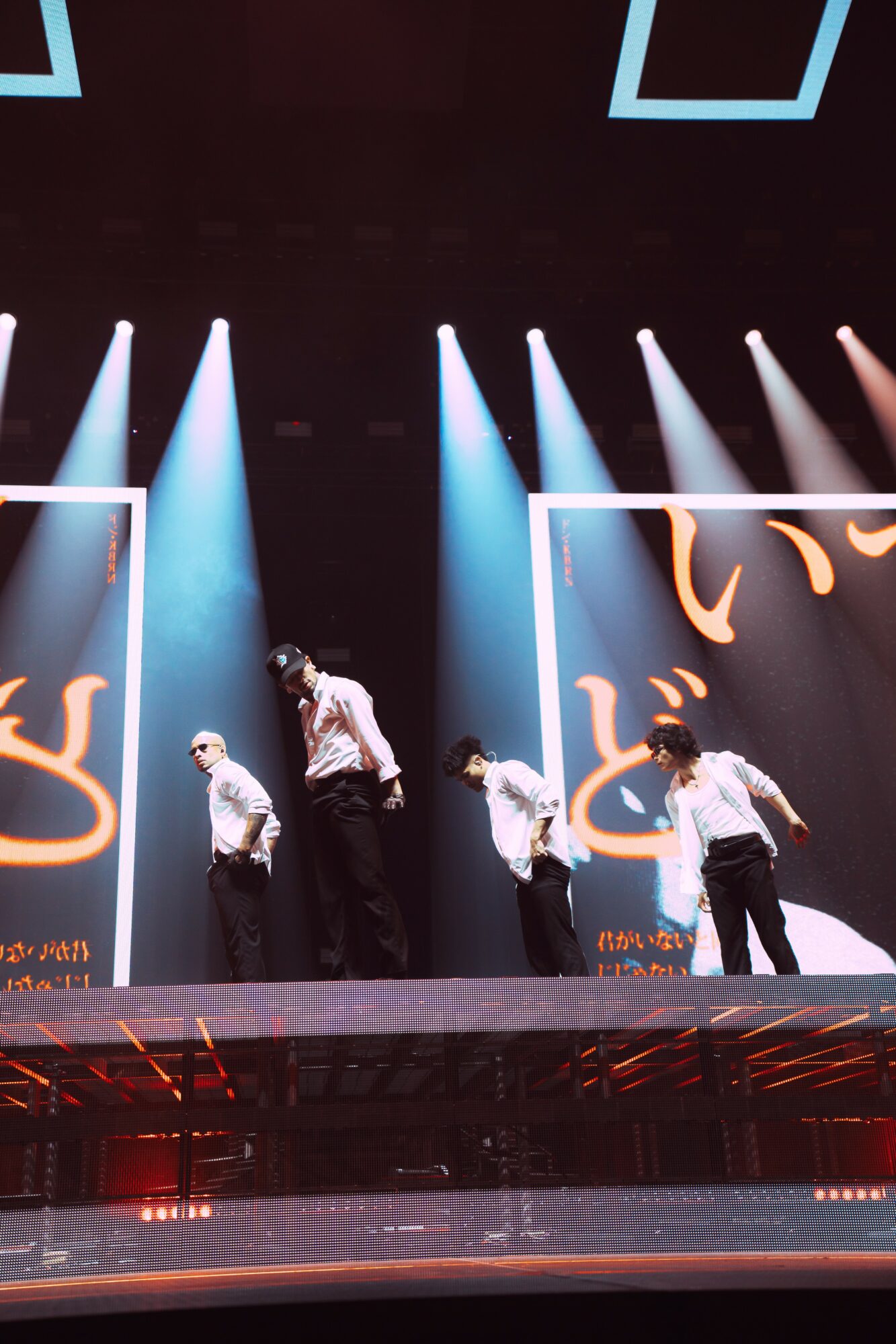 Four performers on stage with blue and white lighting, large screens with abstract designs behind them.