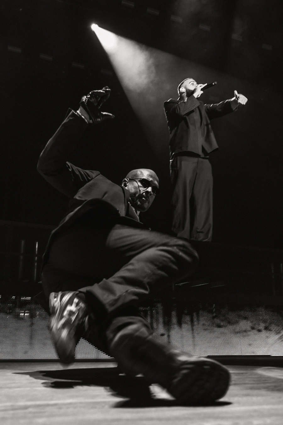 Two performers on stage, one crouching with a mask and the other standing, gesturing with a microphone, spotlight shining down.