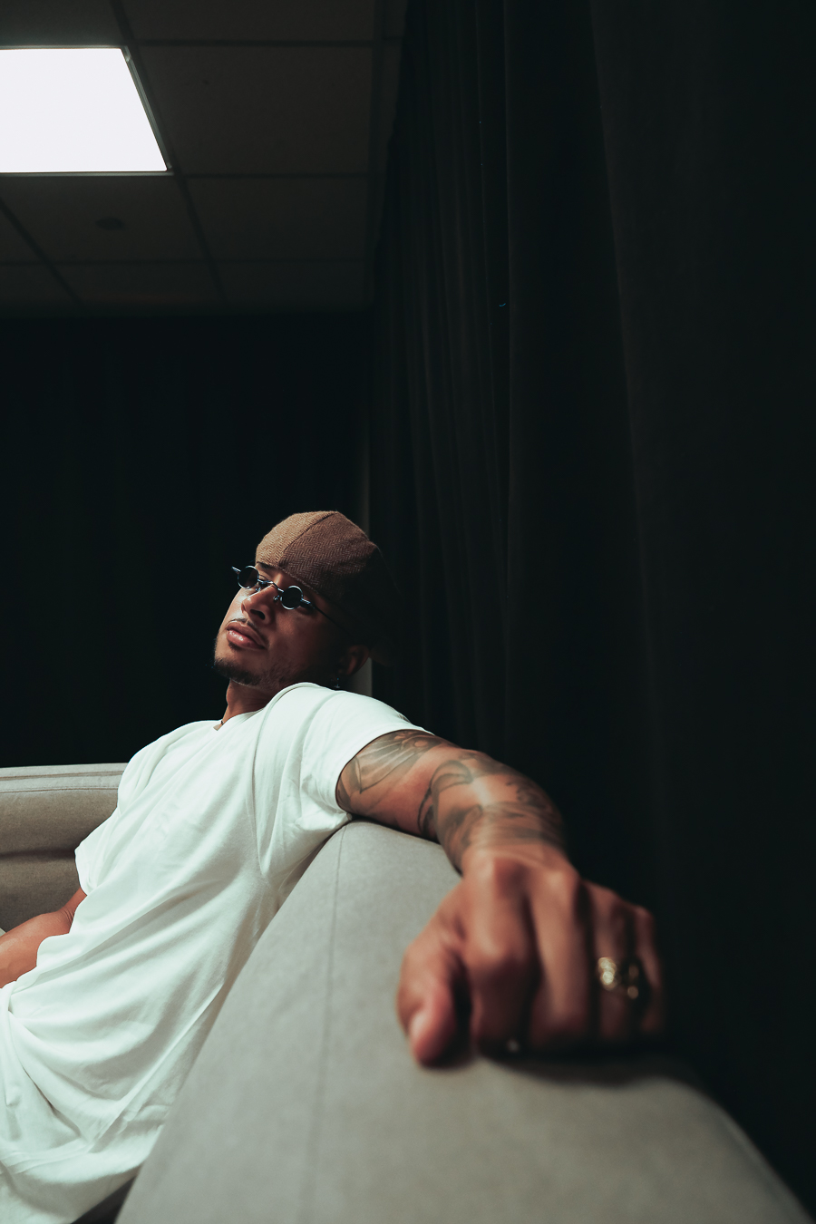 Person with glasses and tattoos sitting on a light-colored couch, arm extended forward, in a dark room with a ceiling light.