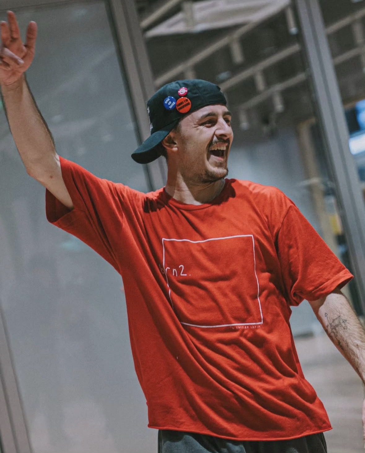 Young man with a beard wearing a red t-shirt and a black cap, smiling and raising his arm, in an indoor setting.