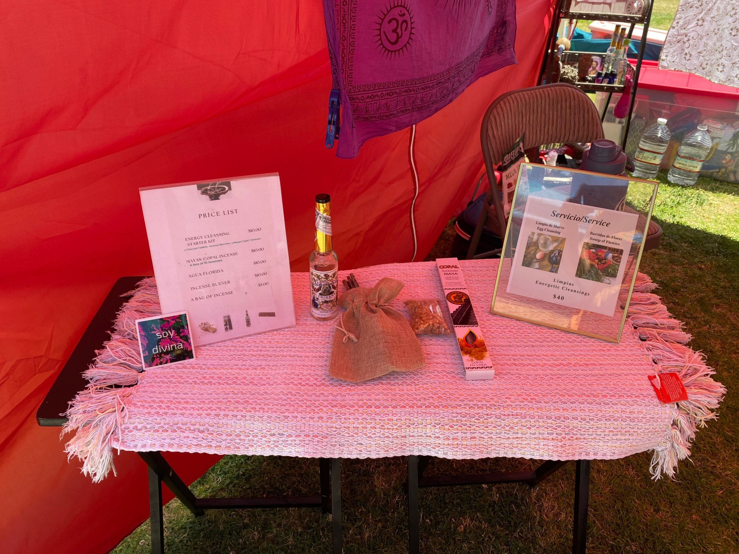 Table with a pink cloth, a chair, a menu, a bottle, and a small bag inside a red tent, outdoors.