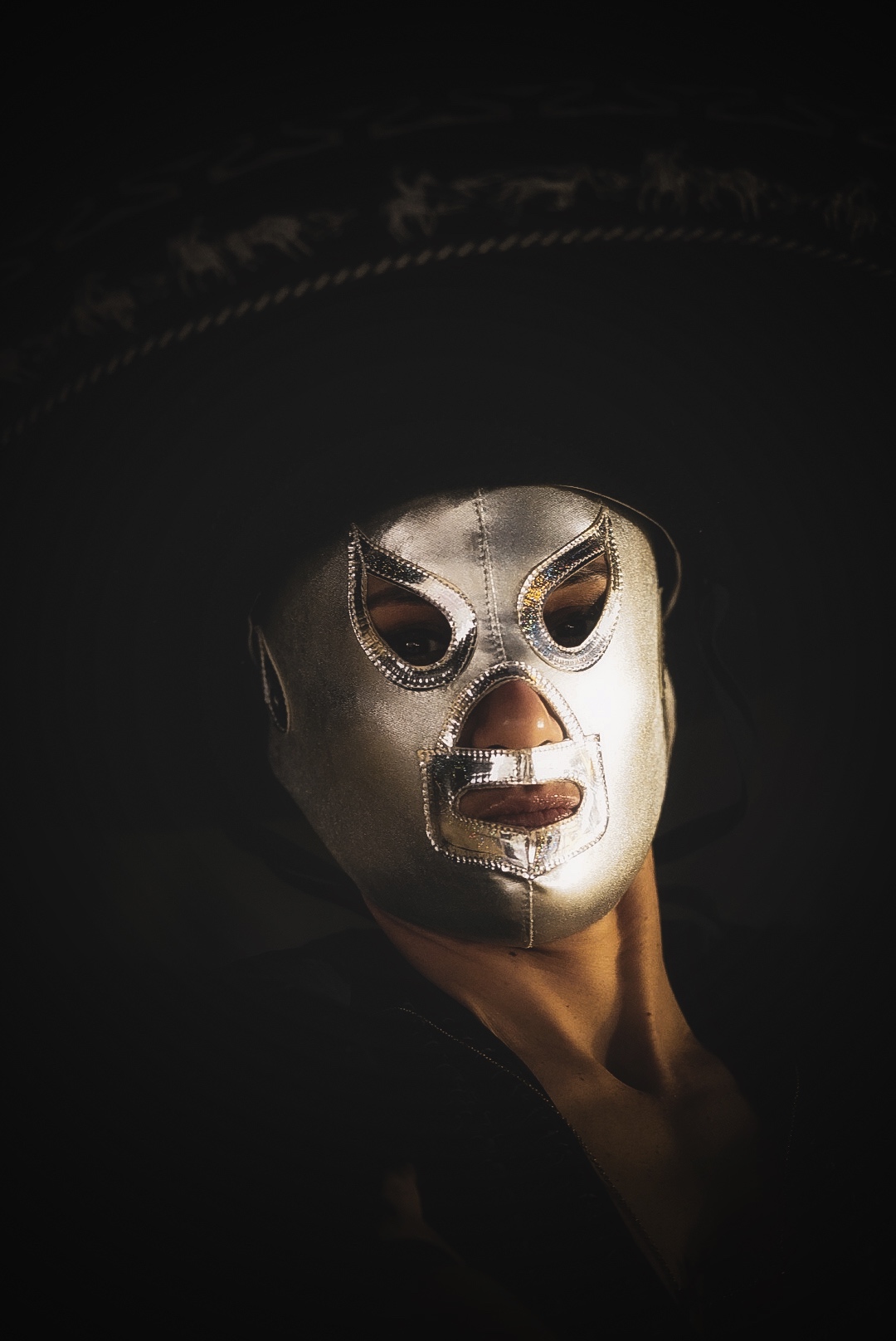 Person wearing a silver mask with dark eye and mouth openings, dark background, face partially illuminated.