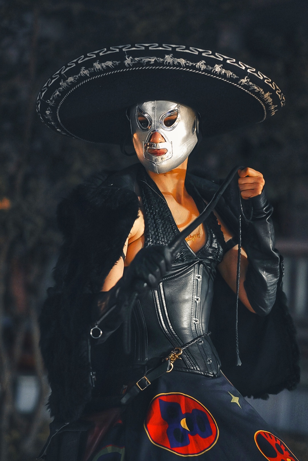 Person wearing a large sombrero, metallic mask, black leather outfit, and holding a whip, with a dark background.