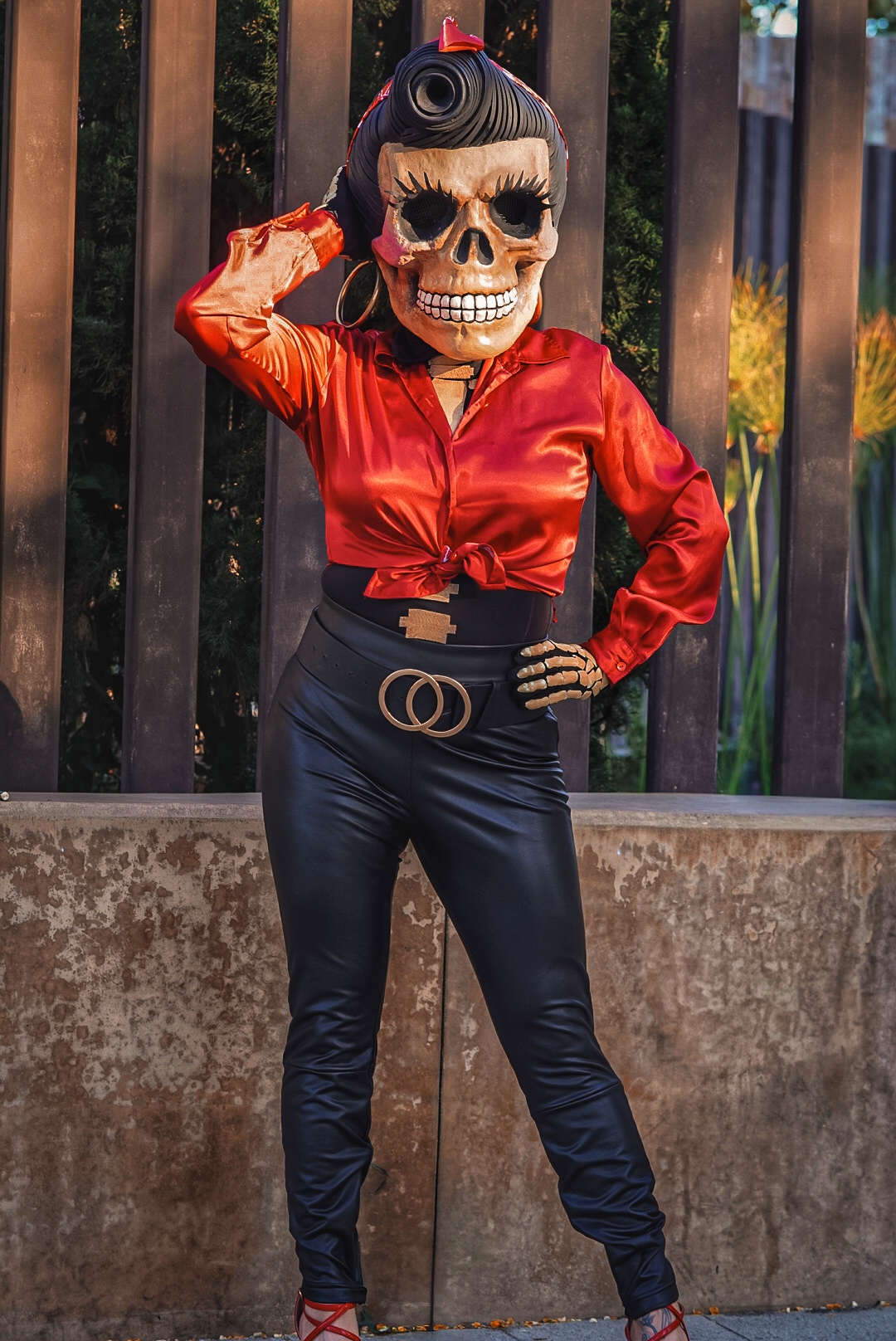 Person wearing a skull mask, red satin blouse, black pants, and standing outdoors with a fence background.