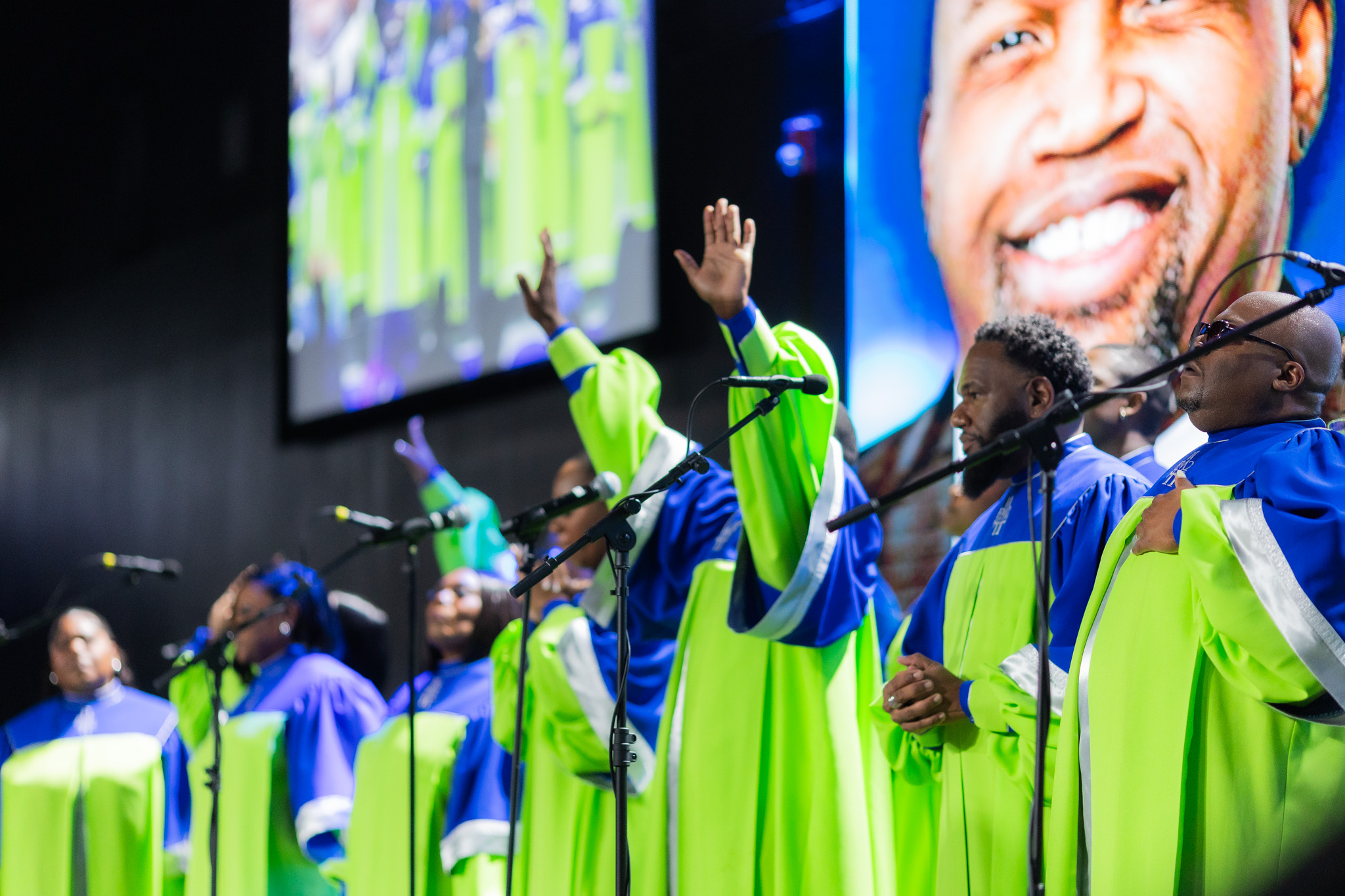 Group of people in colorful robes speaking into microphones on stage, large screen with smiling man's face in background.